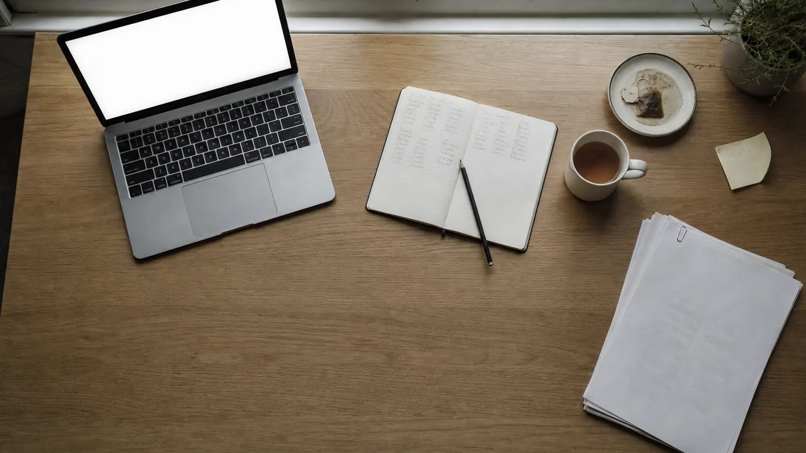 Overhead view of a plain wooden desk by a window: an open laptop, a paper notebook with handwritten columns of figures, a pencil and a mug of tea in soft daylight
